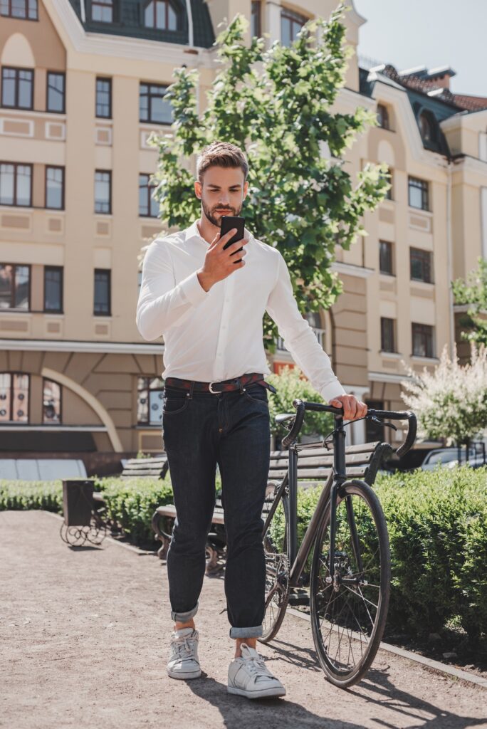 Get busy. Young brown-haired man standing in the park with a bicycle and looking at his phone