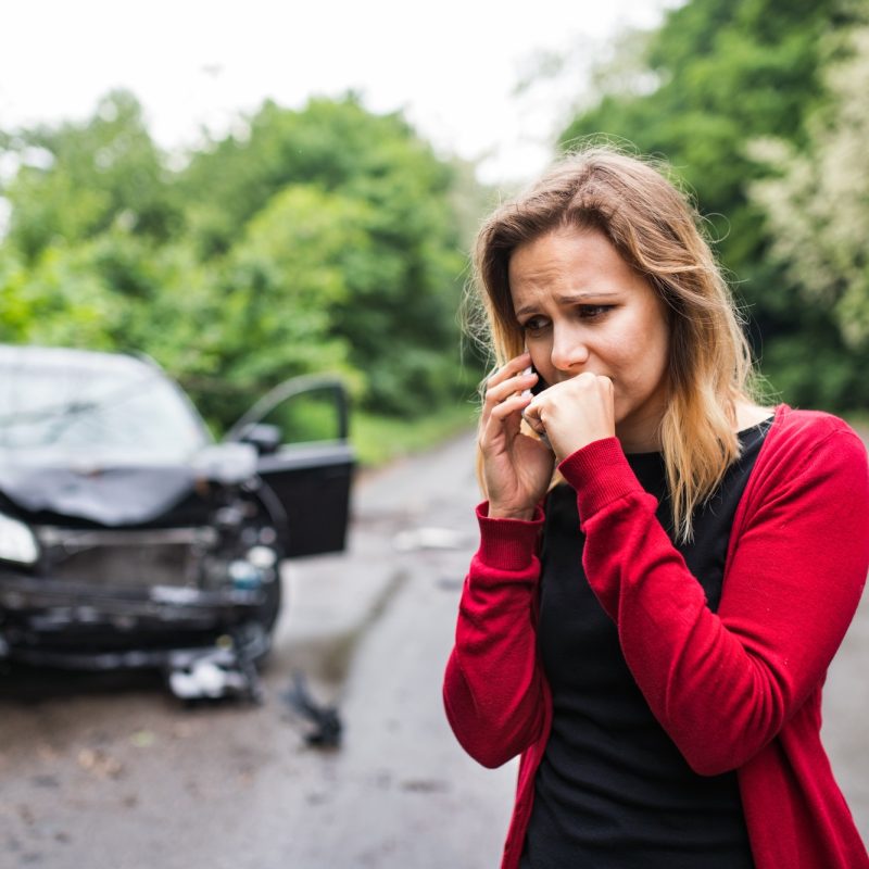 A young woman with smartphone by the damaged car after a car accident, making a phone call.