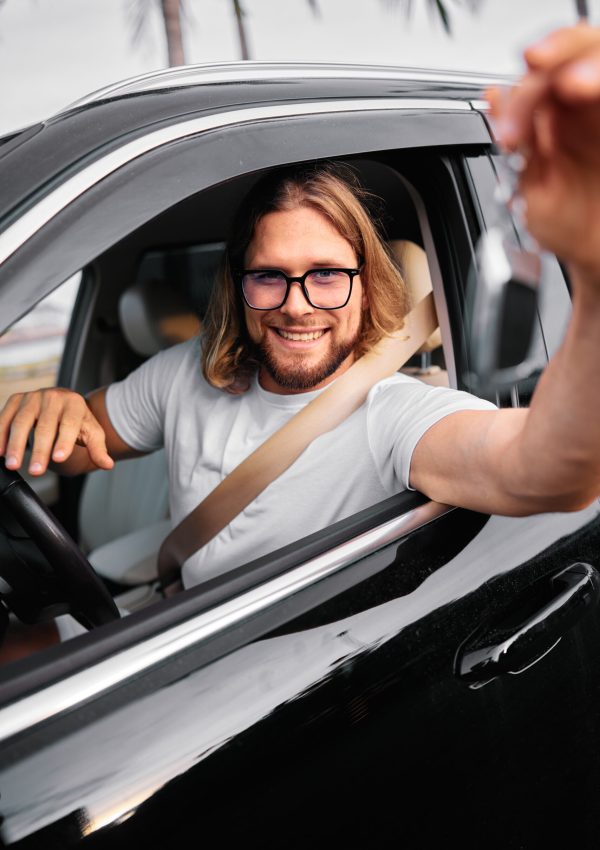 Glücklicher Mann in lässiger Kleidung sitzt lächelnd in einem Auto und hält Autoschlüssel in der Hand Tropische Landschaft in der